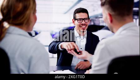 Ein junger Verkäufer lächelt, während er einem Paar, das im Händlerbetrieb ein neues Auto kauft, die Schlüssel übergibt. Stockfoto