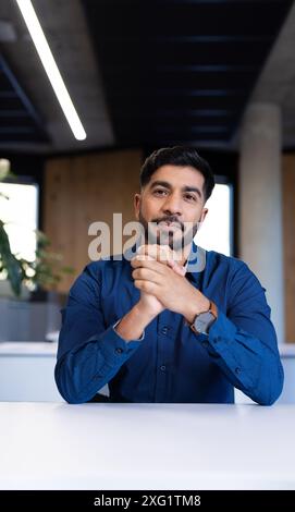 Geschäftsmann in blauem Hemd, der am Schreibtisch sitzt und selbstbewusst in die Kamera und den Kopierraum schaut Stockfoto