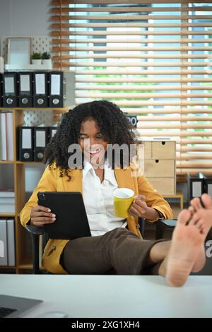 Glückliche afroamerikanische Frau, die sich mit Beinen auf dem Tisch entspannt und ein digitales Tablet verwendet Stockfoto