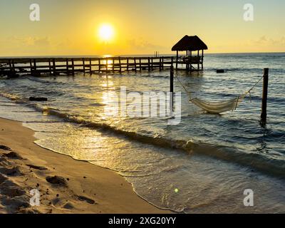 Spektakulärer orangener Sonnenaufgang an einem Strand aus feinem goldenem Sand in der mexikanischen Karibik, mit einem hölzernen Pier und Palapa im Hintergrund über dem Kristall Stockfoto