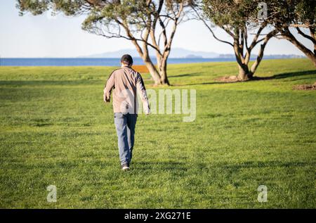 Mann, der auf grünem Gras zwischen Pohutukawa-Bäumen läuft. Rangitoto Island in der Ferne. Milford Beach. Auckland. Stockfoto