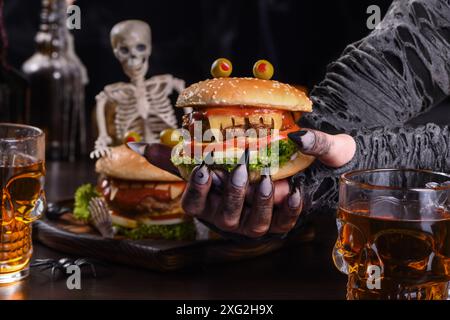 Die Hexenhände halten einen Monster Burger in ihren Händen auf einem sitzenden Skelett. Die perfekte Vorspeise für die Halloween Party Stockfoto