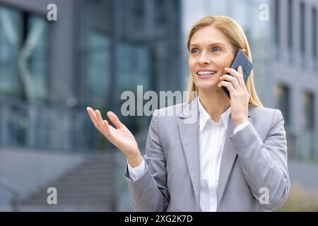 Selbstbewusste Geschäftsfrau in grauem Anzug, die vor einem modernen Bürogebäude steht und ein Telefongespräch führt. Sie lächelt und setzt Gesten ein, während sie über arbeitsbezogene Angelegenheiten spricht. Stockfoto