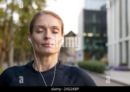 Ruhige Sportlerin beim Ausruhen und Atmen mit Kopfhörern nach einem Workout im Freien, in Sportbekleidung. Sie wirkt entspannt und konzentriert in einer urbanen Umgebung und veranschaulicht Fitness und Wellness. Stockfoto