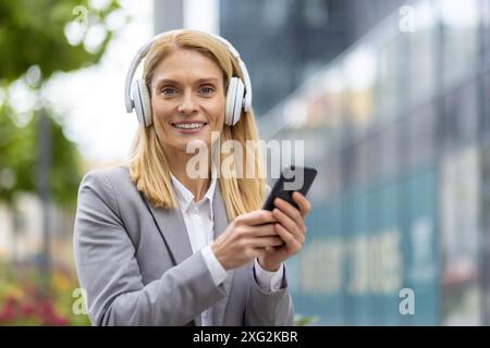 Selbstbewusste und glückliche professionelle Frau, die Kopfhörer trägt und ein Smartphone im Freien in einer urbanen Umgebung hält. Sie ist in Geschäftskleidung gekleidet und steht in einer modernen Stadtumgebung. Stockfoto