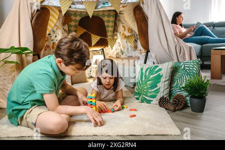 Fröhliche Kinder spielen zusammen mit dem Stapelspiel vor dem hausgemachten Zelt im Wohnzimmer, während die Mutter auf dem Sofa ein Buch liest. S Stockfoto