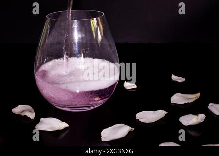 Ein rosafarbenes Getränk wird in ein stielloses Glas gegossen, umgeben von weißen Rosenblättern, Stockfoto