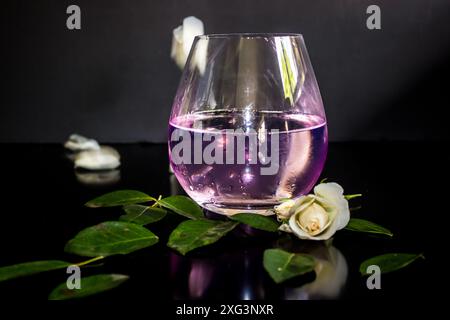 Ein Glas rosa Gin Tonic, umgeben von Rose, Blumen und Blättern, mit fallenden Blütenblättern im Hintergrund Stockfoto