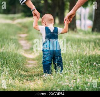 Eltern halten die Hände ihres Babys, während sie auf einem grasbewachsenen Weg nach draußen gehen, während das Kind blaue Overalls trägt. Die Szene fängt einen zarten Moment ein Stockfoto