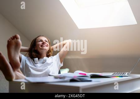 Bildung und sorgloser Student. Junge Frau mit Füßen auf dem Tisch und Händen hinter dem Kopf sitzt zu Hause am Tisch und lächelt. Stockfoto