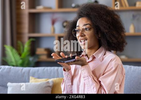 Junge Frau, die mit der Spracherkennungsfunktion in das Smartphone spricht. Sie sitzt auf einer Couch in einer entspannten Wohnumgebung und trägt eine Brille, die verlobt aussieht. Moderne Technologie und Kommunikation Stockfoto