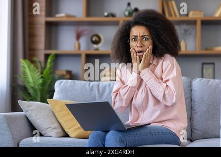 Schockierte Frau mit einem Laptop, die zu Hause mit einem überraschenden Ausdruck auf einem Sofa sitzt. Stellt Konzepte von unerwarteten Ereignissen, Überraschungen, schockierenden Nachrichten und emotionalen Reaktionen dar Stockfoto