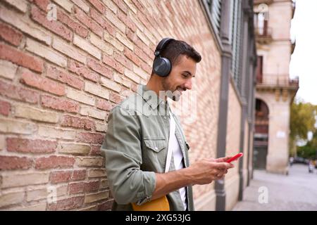 Attraktiver junger kaukasischer Mann mit schnurlosen Kopfhörern, der ein Handy gegen eine Ziegelwand lehnt. Stockfoto