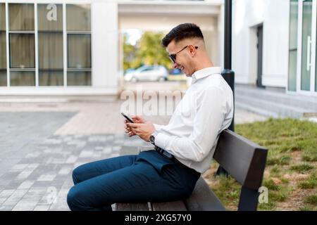Ein Mann sitzt draußen auf einer Bank, vertieft in sein Handy. Er scheint auf dem Bildschirm konzentriert zu sein, möglicherweise SMS zu schreiben oder zu stöbern. Stockfoto