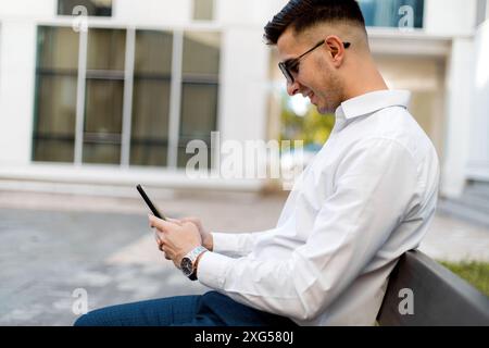 Ein Mann sitzt auf einer Bank, vertieft in sein Handy. Er scheint konzentriert zu sein, während er durch den Bildschirm blättert, ohne seine Umgebung zu vergessen. Stockfoto