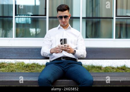 Ein Mann sitzt auf einer Bank, vertieft in sein Handy. Er scheint auf dem Bildschirm konzentriert zu sein, wobei das Gerät in beiden Händen gehalten wird. Stockfoto