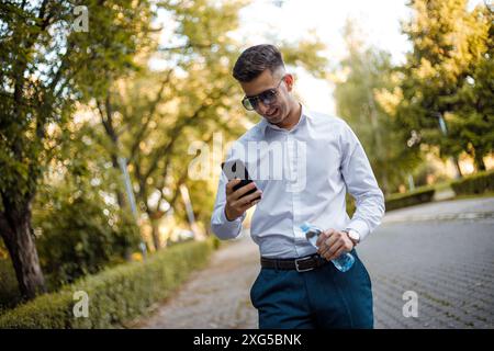 Ein Mann in einem weißen Hemd und einer blauen Hose steht und blickt aufmerksam auf ein Handy in seinen Händen. Stockfoto