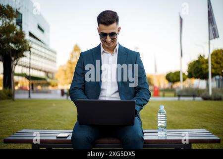 Ein Mann sitzt draußen auf einer Holzbank und arbeitet an seinem Laptop. Er konzentriert sich auf den Bildschirm und auf das Tippen. Stockfoto