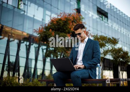 Ein Mann sitzt auf einer Bank in einem öffentlichen Bereich, vertieft in seine Arbeit auf einem Laptop. Er scheint auf dem Bildschirm konzentriert zu sein. Stockfoto