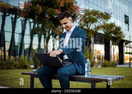 Ein Mann in einem formellen Anzug sitzt draußen auf einer Bank. Er konzentriert sich auf seinen Laptop-Bildschirm, möglicherweise arbeitet oder studiert. Stockfoto