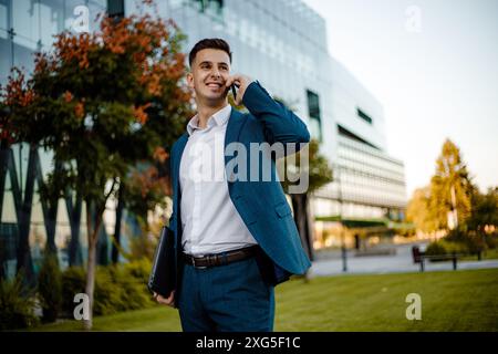 Ein junger Mann in blauem Anzug und weißem Hemd steht vor einem Bürogebäude, hält einen Laptop und spricht mit seinem Handy. Stockfoto