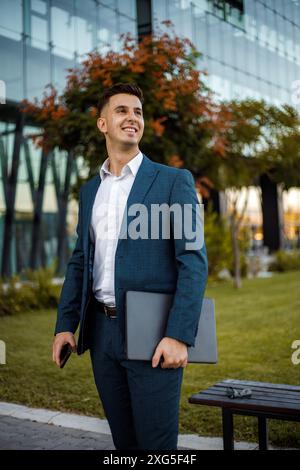 Ein junger Geschäftsmann in einem blauen Anzug steht vor einem modernen Bürogebäude und hält einen Laptop in der Hand. Stockfoto