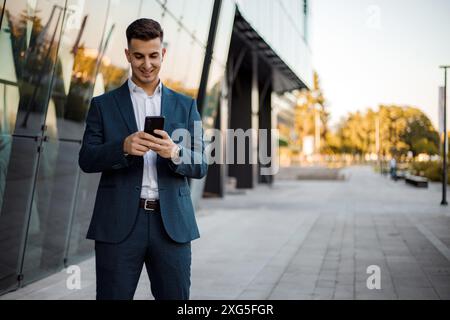 Ein junger Mann im Anzug steht an einem sonnigen Tag auf dem Bürgersteig vor einem Bürogebäude. Er blickt mit einem Lächeln auf sein Smartphone. Stockfoto