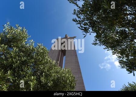 Niedriger Blick auf die Statue Cristo Rei, eingerahmt von Bäumen unter blauem Himmel - Almada, Lissabon, Portugal Stockfoto