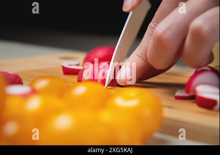 Nahaufnahme weiblicher Hände Schnitt frischen rohen Rettich auf einem Holzbrett vor dem Hintergrund gelber Kirschtomaten. Vegetarisches leckeres und gesundes Essen Stockfoto