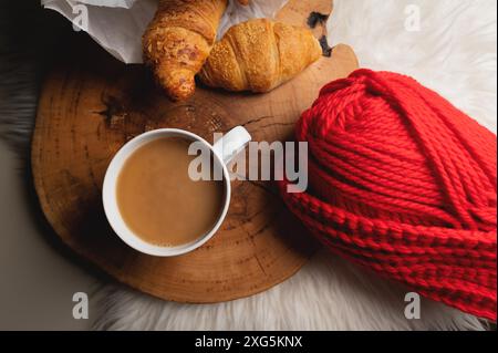 Stimmungsvolle Stilleben weiße Tasse Kaffee neben frischen Croissants in einer Papiertüte neben rotem Garn. Komfort für Ihr Zuhause mit weichem Fokus. Draufsicht Stockfoto
