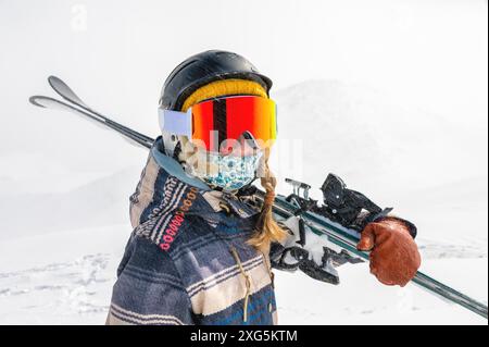 Eine junge Frau im Skiurlaub, steht und hält Skier auf der Schulter vor der Kulisse der Berge. Abseits der Pisten, Freeride Stockfoto