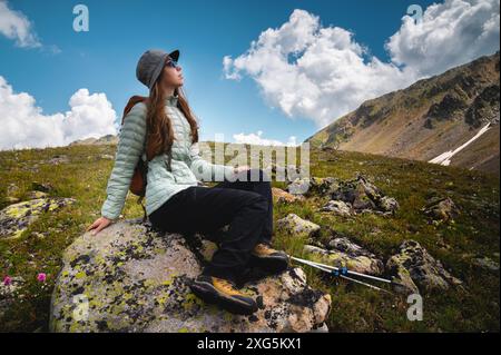 Eine junge Frau mit Rucksack sitzt auf einem Stein und schaut in den Himmel mit Wolken und Bergen, Wanderurlaub Stockfoto
