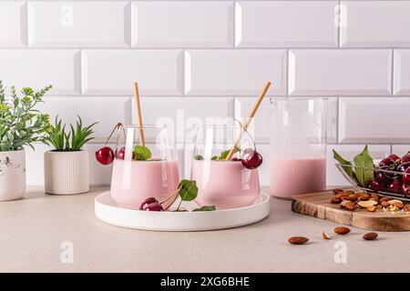 Köstlicher gesunder Kirsch-Smoothie in zwei Glasgläsern mit Öko-Stroh auf einem Keramiktablett auf einer Steinplatte mit Kochzutaten. Stockfoto