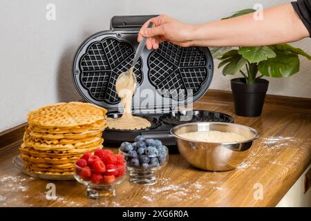 Belgische Waffeln machen. Weibliche Hand gießt Teig in die Waffelmaschine in der Küche zu Hause. Kochendes Sommerfrühstück mit runden Waffeln und Beeren Stockfoto