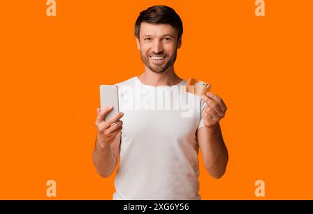 Mann hält Telefon und Kreditkarte vor orangefarbenem Hintergrund Stockfoto