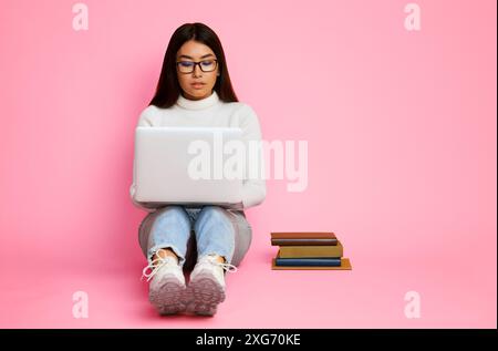 Junge Frau benutzt Laptop, während sie auf rosa Hintergrund sitzt Stockfoto