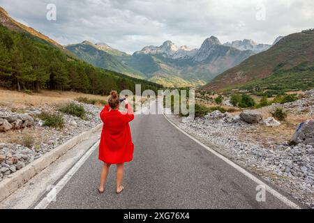 Frau, die ein Foto von einer Bergstraße macht, mit einem Kameratelefon in der Hand und einer Bergkette im Hintergrund, Albanien, in der Nähe des Theth-Tals Stockfoto