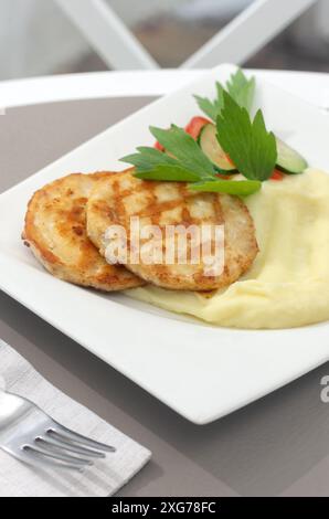 Zwei knusprige Fleisch- oder Hühnchenkoteletts in goldener Panierung, serviert mit Kartoffelpüree, frischem Dill, Kirschtomaten auf einer weißen Keramikplatte. Auf einer Leuchte bac Stockfoto