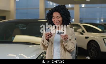 Afroamerikaner glücklicher Kunde weibliche Käuferin halten Kreditkarte Handy Shopping kaufen neue Luxus-Elektroauto im Autohaus des Autohauses Stockfoto