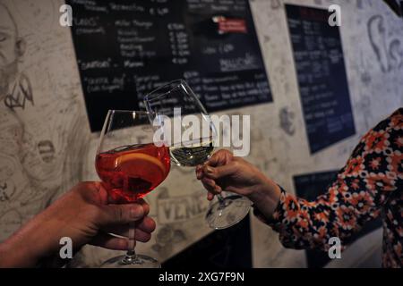 Zwei Hände mit Gläsern Campari Spritz und Prosecco Wein in einem kleinen bácaro mit Graffiti-Kunstwand und Tafel-Getränkekarte in Venedig Stockfoto