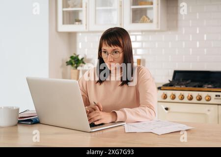 Ernsthafte Frau, die eine Brille trägt und online an einem Laptop arbeitet, am Tisch in der Küche sitzt und auf den Computerbildschirm blickt. Fokussierte Frau, die das Internet für Fernunterricht nutzt, Informationen sucht, Fähigkeiten verbessert Stockfoto