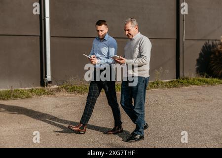 Zwei Männer gehen Seite an Seite auf einem gepflasterten Weg außerhalb eines großen Gebäudes, beide tragen legere Kleidung und halten Tablets. Stockfoto