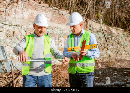 Zwei Bauarbeiter mit Schutzhüten und Sicherheitswesten arbeiten an einem Bauprojekt. Ein Arbeiter hält ein Maßband und verlängert sich Stockfoto