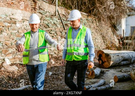 Zwei Bauarbeiter in Schutzhelmen und Sicherheitswesten messen auf einer Baustelle einen langen, dünnen Metallstab. Es gibt verschiedene d Stockfoto