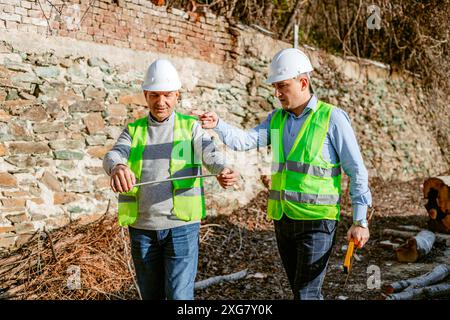 Zwei Bauarbeiter, die weiße Schutzhelme und hellgrüne Sicherheitswesten tragen, stehen vor einer Felswand. Stockfoto