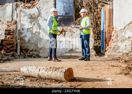 Zwei Männer mit Schutzhüten und reflektierenden Westen stehen vor einem teilweise abgerissenen Gebäude und diskutieren anscheinend Pläne für das Bauprojekt. Stockfoto