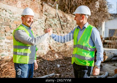 Zwei Bauarbeiter, die weiße Schutzhelme und gelbe Sicherheitswesten tragen, stehen vor einer Ziegelmauer und schütteln die Hände. Stockfoto