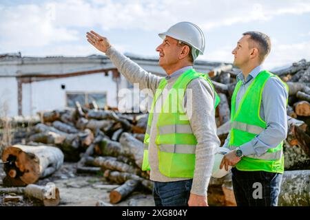 Zwei Bauarbeiter, einer mit Schutzhelm, stehen neben einem großen Holzhaufen. Sie schauen sich etwas in der Ferne an und reden mit ihm Stockfoto