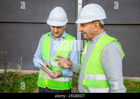 Zwei Bauarbeiter mit Schutzhüten und Sicherheitswesten stehen tagsüber vor einem Gebäude. Stockfoto