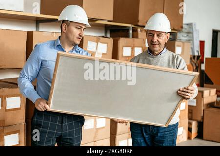 Zwei Männer mit Schutzhüten halten eine große, gerahmte Leinwand in einem Lagerhaus. Sie stehen vor Regalen, die mit Kartons gestapelt sind. Stockfoto